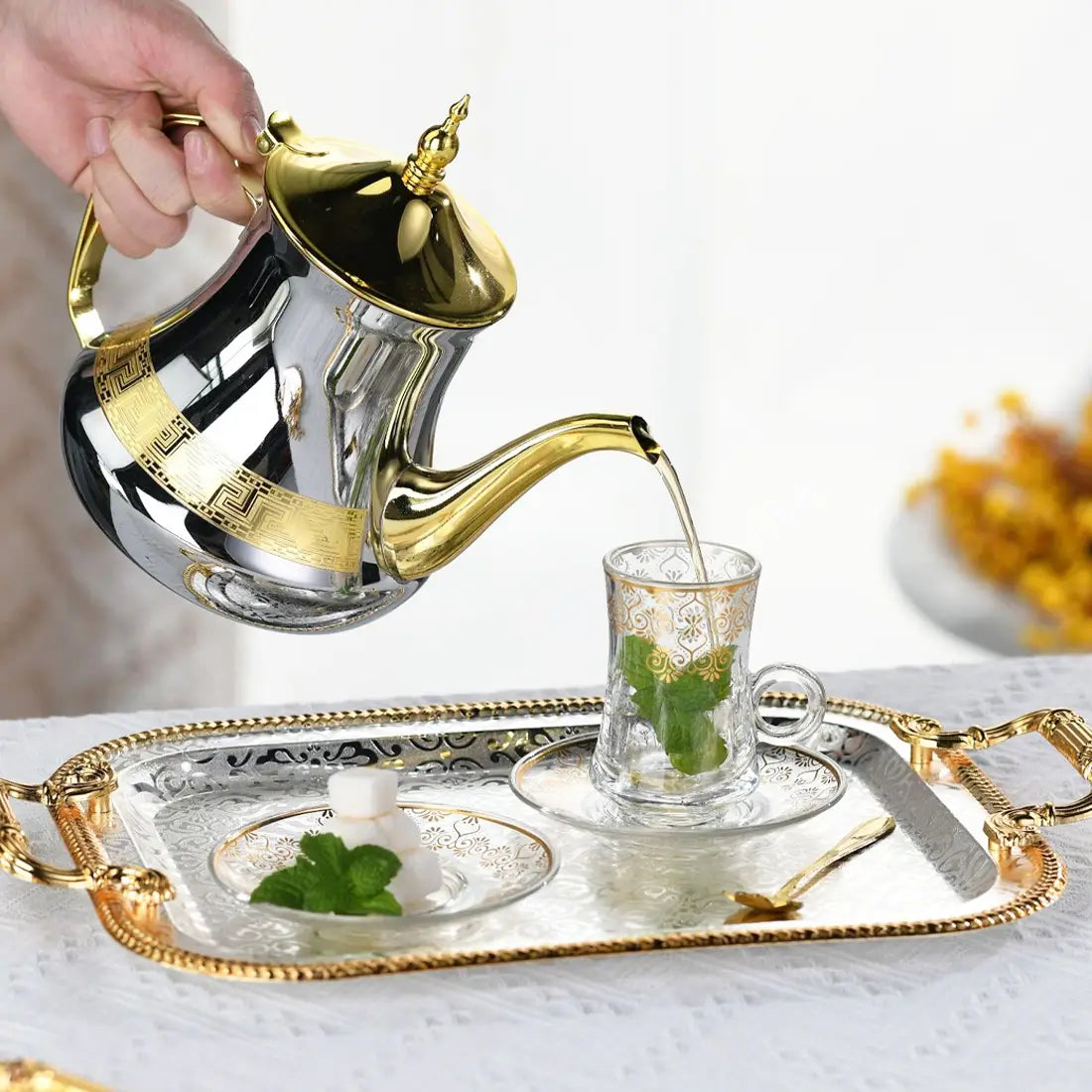 A luxury rectangle serving tray with gold handles holds a gleaming gold and silver teapot featuring intricate geometric patterns pouring tea into a matching glass cup.