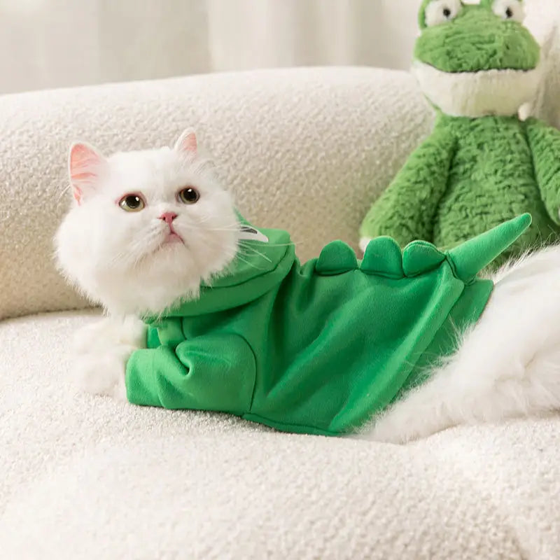 A fluffy white cat wearing a vibrant green dinosaur cat hoodie made of plush material featuring a playful spiked tail design.