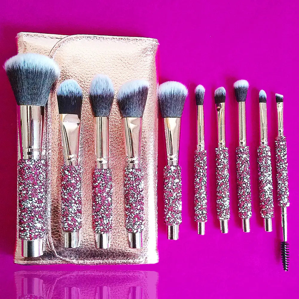 Set of ten glittery pink and silver rhinestone makeup brushes with metallic handles neatly arranged in a rose gold textured pouch.