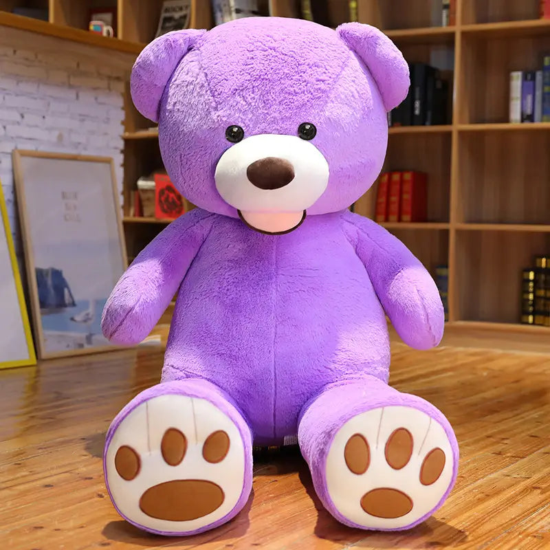 Ein großer Teddybär-Plüschartikel aus violettem Samt mit weißen Pfotenpolstern und braunen Zehen-Details, der auf einem Holzboden sitzt.