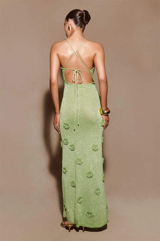 A back view of a light green crochet floral lace mermaid maxi dress with glitter accents featuring a backless design with crisscross straps and decorative rose appliqués.
