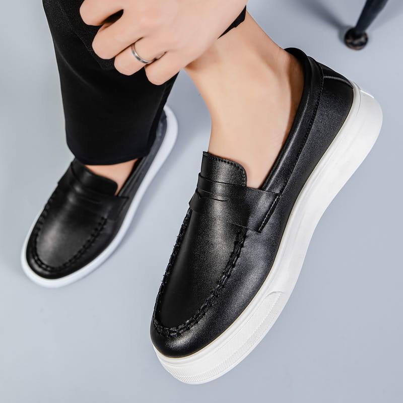 Mocasines negros de cuero, tipo slip-on, con suela de goma blanca y detalles cosidos.