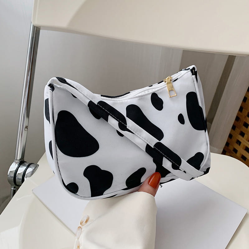 A white and black cow print crossbody bag with a gold zipper designed as an animal print underarm shoulder bag for women.