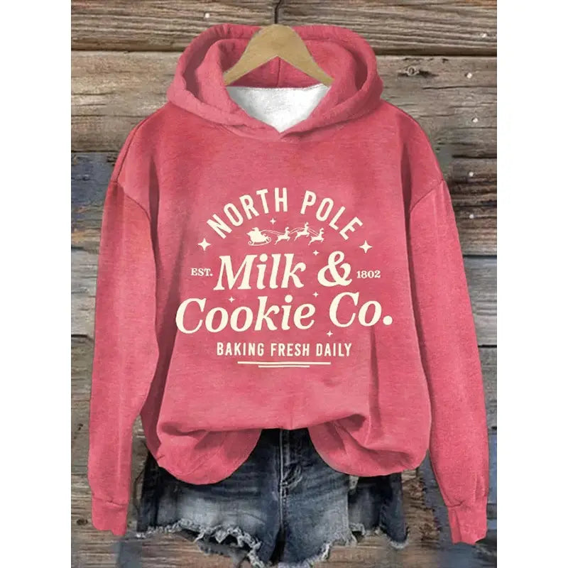 Sudadera con capucha rosa suave con un gráfico de leche y galleta de polo norte blanco y un diseño festivo de renos, colgada de una percha de madera titulada sudadera con capucha de estampado de olas japonesas.