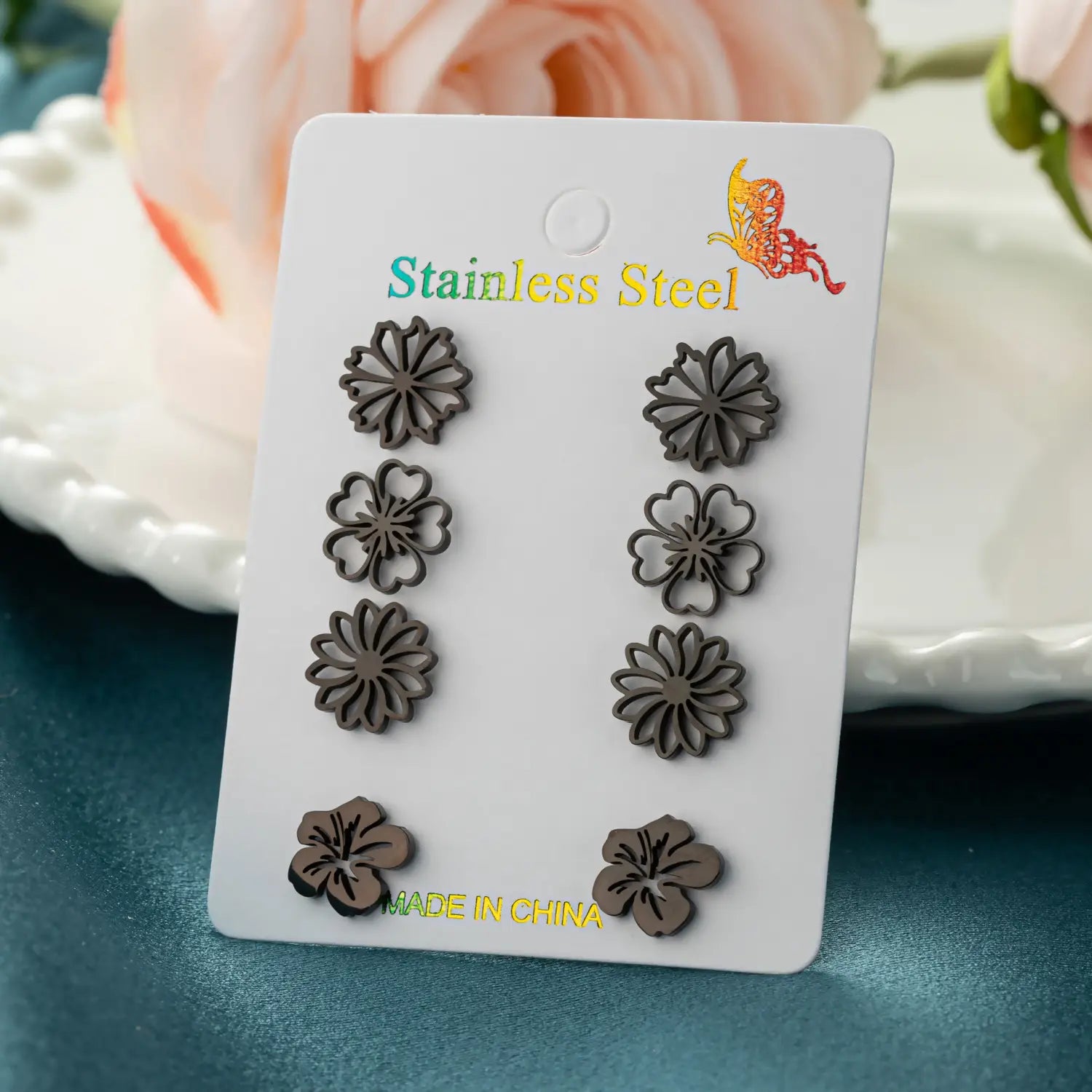 Ensemble de six boucles d’oreilles à clous en acier inoxydable à motif floral de couleur gris foncé, présentées sur une carte blanche.