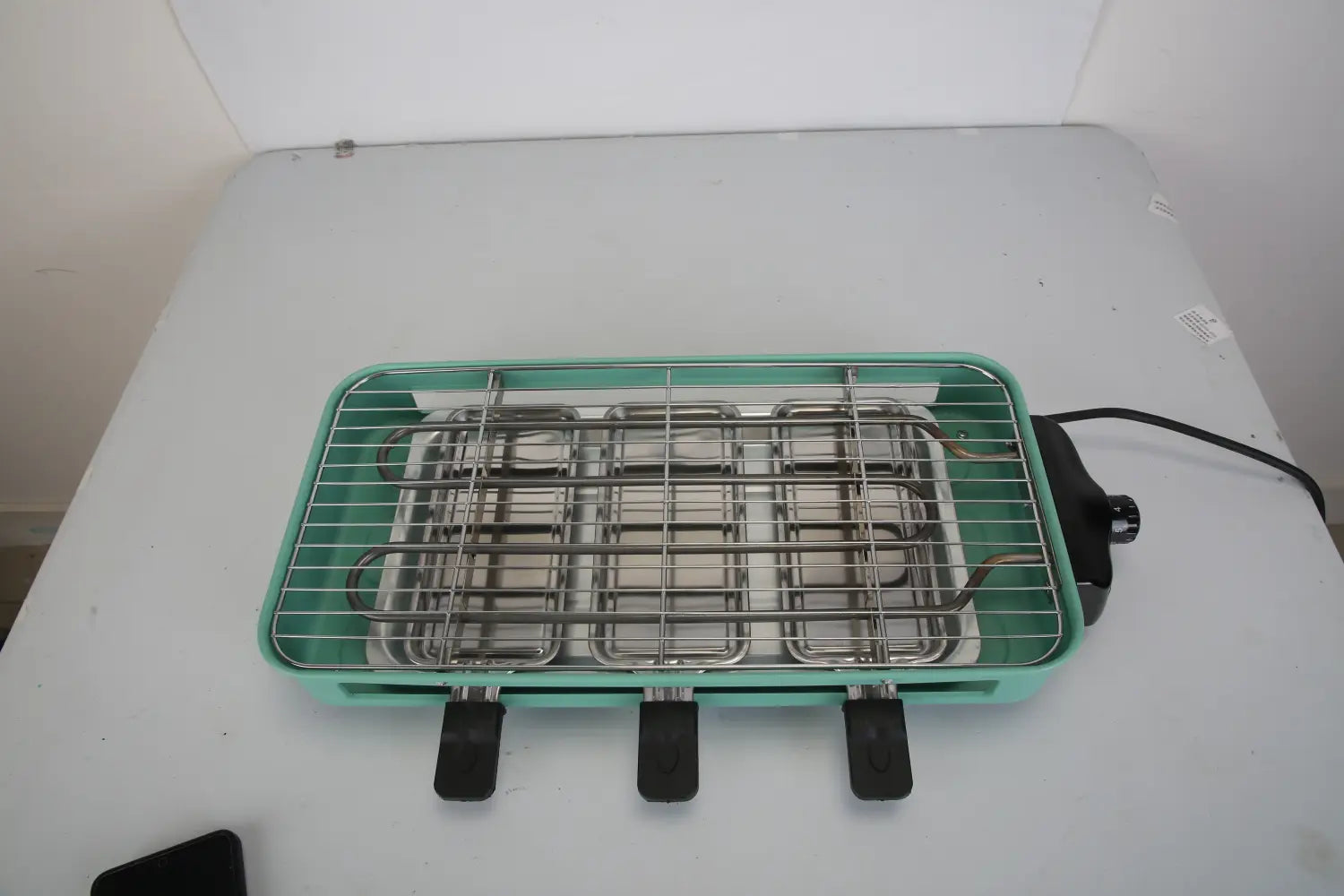 Gril électrique intérieur de 2000 W de couleur bleu-vert, avec une grille en fil métallique en métal et trois poignées noires, comprenant trois bacs à raclette pour une cuisson polyvalente.