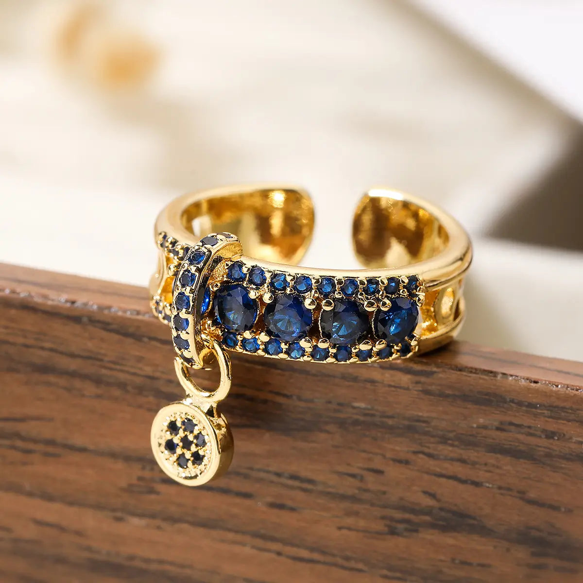 Adjustable geometric gold open ring featuring a gold-toned band with deep blue zircon gemstones and a dangling circular charm with black accents.