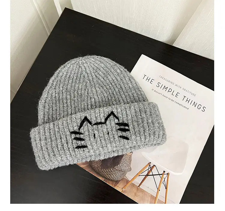 Gray knitted beanie hat with black embroidered cat face on the folded brim designed for warmth and style.