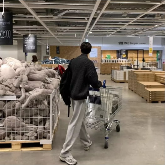 A person wearing a black men’s ribbed knit zip up sweater cardigan and light gray sweatpants pushes a metal shopping cart through an Ikea store aisle.