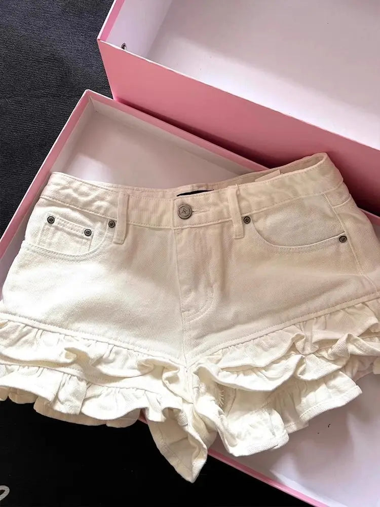 Los pantalones cortos de mezclilla con volantes en color crema presentan un diseño de cintura alta con material de mezclilla color crema y dobladillo con capas de volantes, asegurado mediante un cierre de botones de tono plateado.