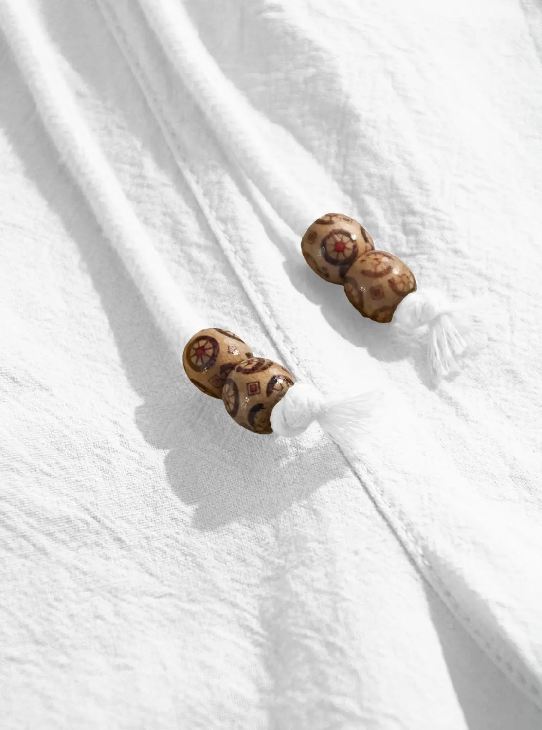 mens cotton linen drawstring shorts with hand-carved wooden beads featuring intricate circular and geometric patterns in warm brown tones strung on white cord.