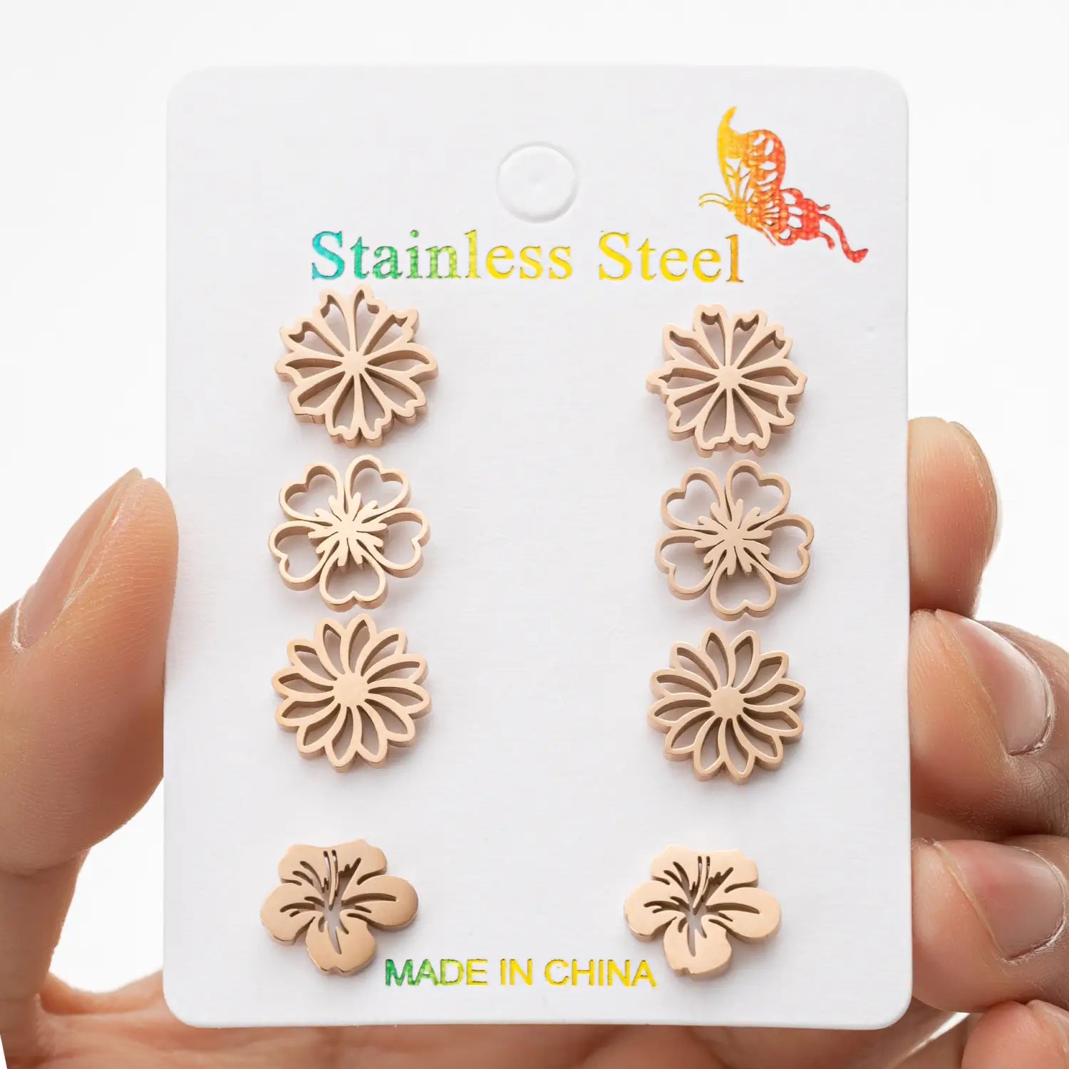 Ensemble de six boucles d’oreilles à clous en acier inoxydable de couleur beige clair, à motif floral, finement découpées et présentées sur une carte blanche.