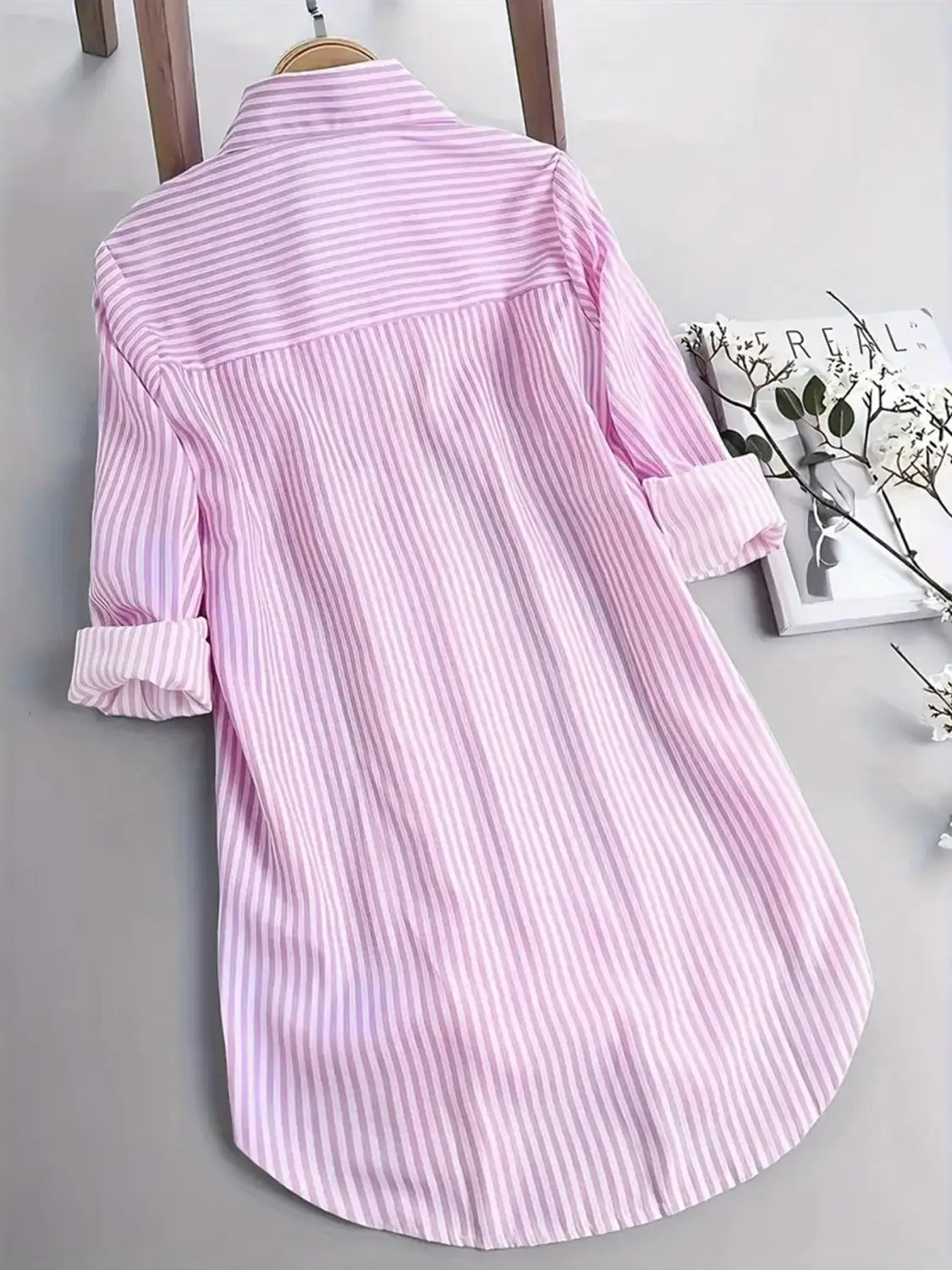 Chemise boutonnée pour femme à manches longues à rayures verticales rose tendre et blanches, poignets retroussés, suspendue à un cintre en bois, adaptée comme haut décontracté.