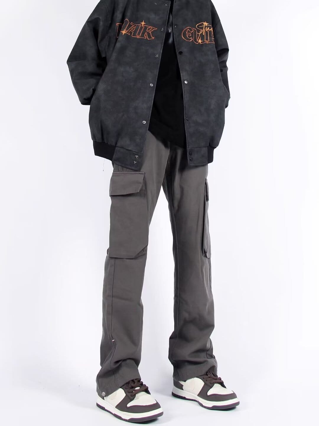 Chaqueta bomber negra con letras bordadas «ak» y «civil» en hilo naranja y dorado, combinada con pantalones cargo rectos de pierna recta con múltiples bolsillos