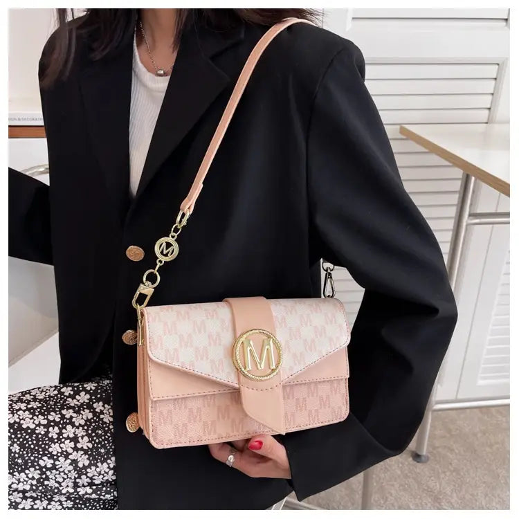 A blush pink monogram letter print crossbody messenger bag with a repeating pattern and gold tone hardware featuring an envelope flap closure.