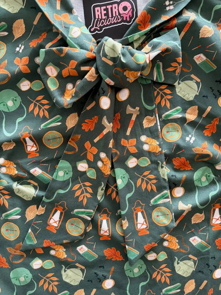 A dark green cotton bow top with vintage camping print featuring orange leaves lanterns and teapots