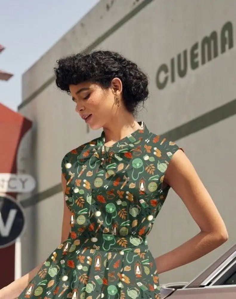 A woman wearing a vintage camping print cotton bow top in dark green with teapots, lanterns, and leaves pattern