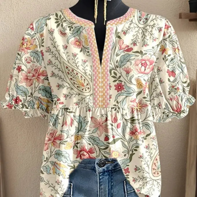 クリームを基調とし、ピンクとグリーンのフローラルおよびペイズリーの模様をあしらったフローラルのVネックラッフルスリーブのブラウスで、ネックラインに繊細なピンクとベージュのチェック仕立てのトリムが施されています。