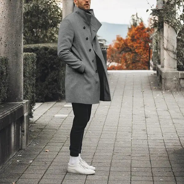 A man wearing a gray double-breasted wool tweed trench coat with black buttons and a stand collar paired with black pants and white sneakers.