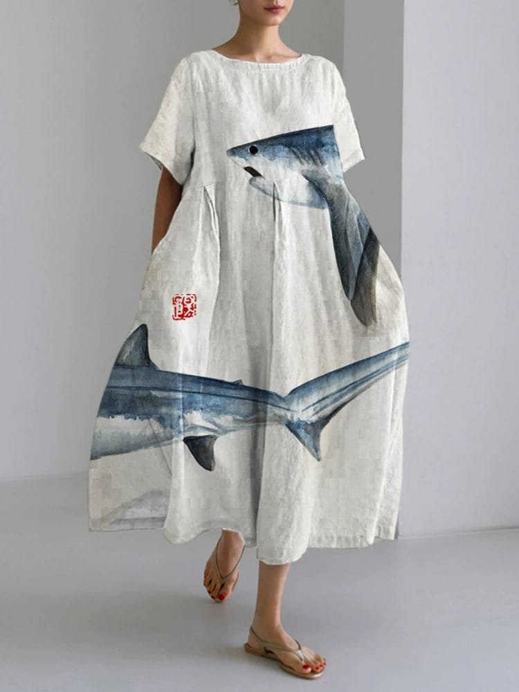 A flowing off-white linen maxi dress featuring a large watercolor-style shark graphic in blue and gray with a small red seal-like emblem on the side pocket.