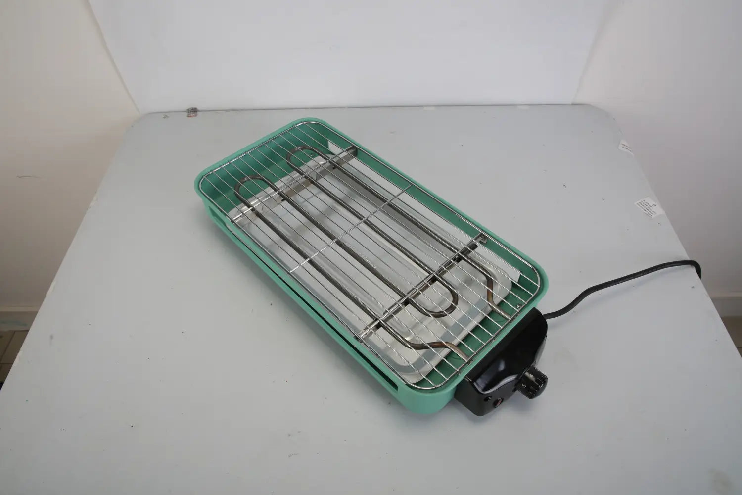 Un gril électrique intérieur de 2000 W de couleur bleu-vert, doté d’un élément chauffant en métal et d’une grille en fil métallique, comprenant trois bacs à raclette pour une cuisson polyvalente.