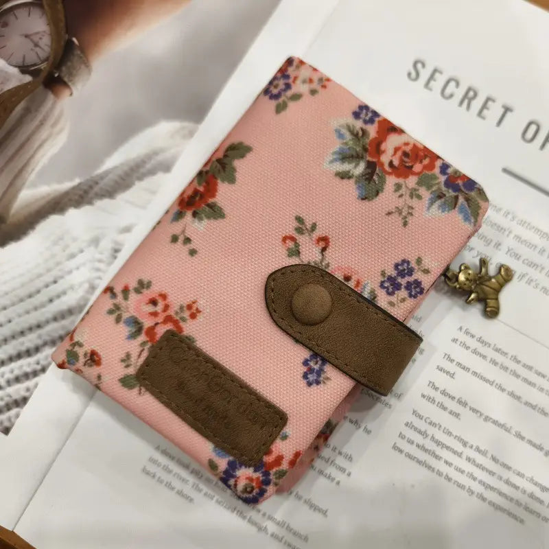 Pink floral-patterned fabric tri fold multi card wallet with brown leather strap and button closure showcasing whimsical patterned canvas design.