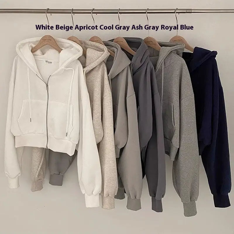 Eine Reihe gemütlicher, verkürzter Zip-up-Kapuzenpullover in weichen Pastell- und neutralen Tönen auf hölzernen Kleiderbügeln, mit einem Design im Stil eines dunkelblauen Samt-Velours, wattierten Kapuzenpullovers mit schmaler Passform