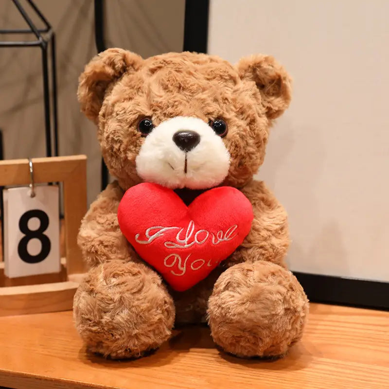 Ein brauner Plüsch-Teddybär mit weißer Schnauze und schwarzen Augen, der ein rotes, mit „I love you“ besticktes Herz hält; geeignet als Valentinsgeschenk in Größen 16 bis 25 cm.
