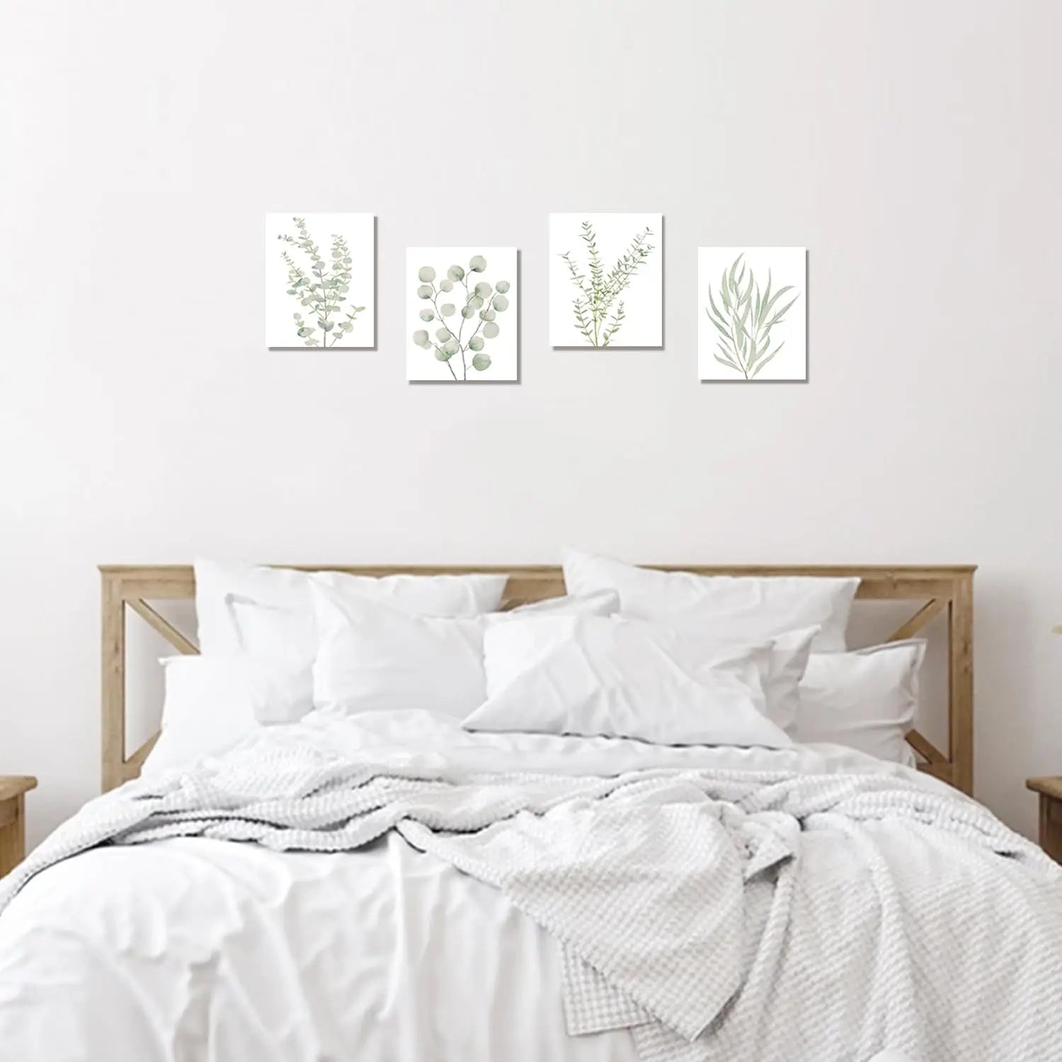 Set of four minimalist botanical canvas wall art prints featuring delicate green and gray eucalyptus foliage on white backgrounds framed and symmetrically arranged above a wooden bed frame.
