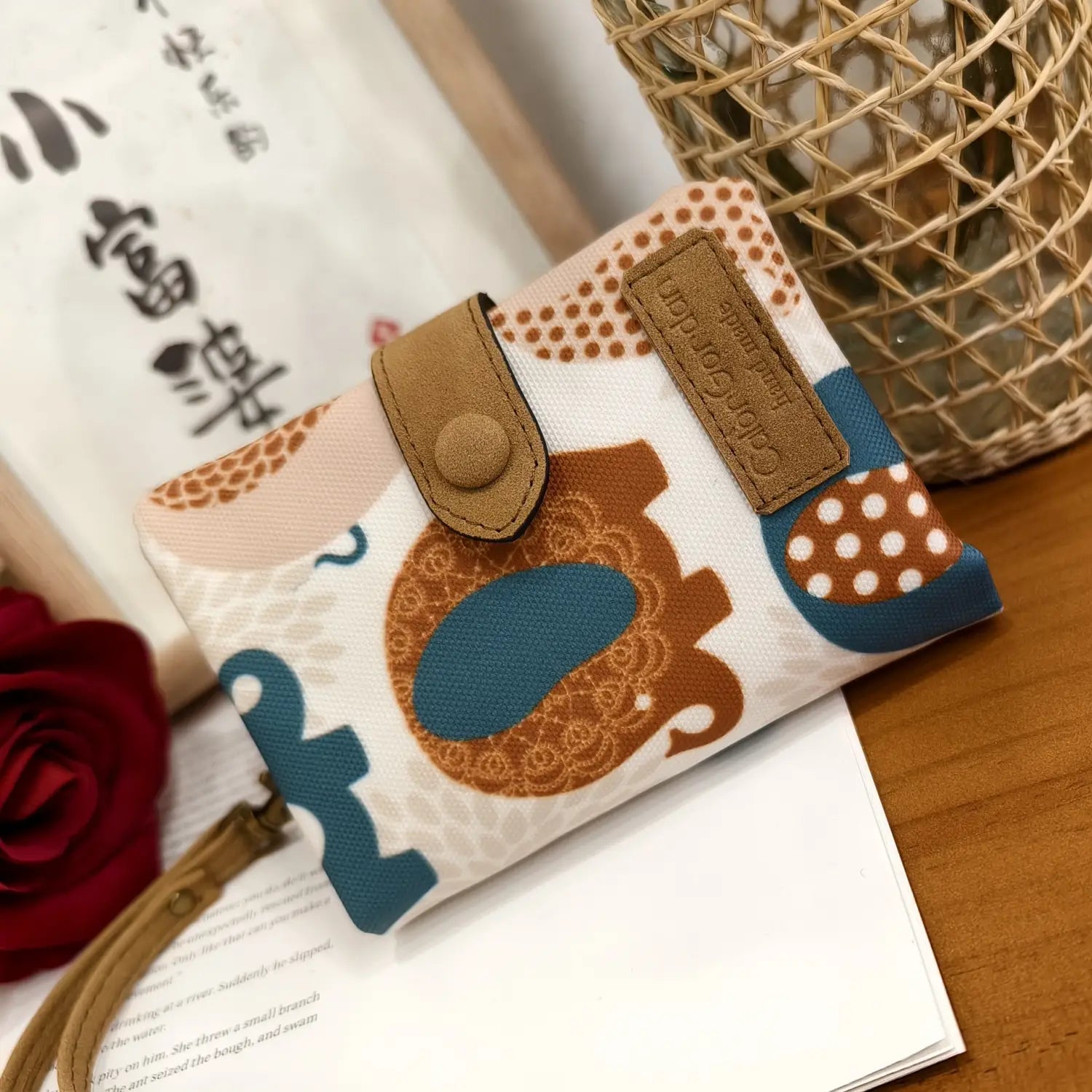 A whimsical patterned beige canvas tri fold multi card wallet featuring a playful elephant design in brown and teal polka dots with tan leather straps and a branded patch.