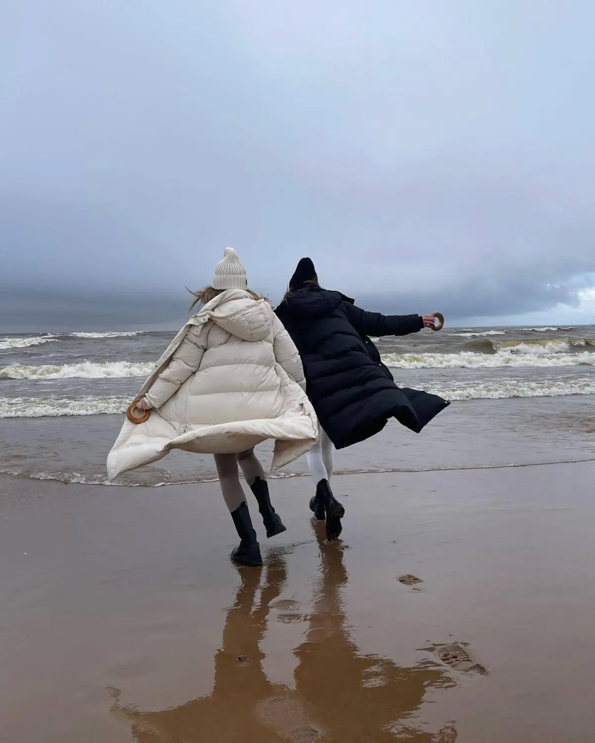 Deux personnes portant des manteaux d’hiver matelassés à capuche, en crème et en noir, courent le long d’une plage humide, avec des vagues qui s’écrasent derrière elles.