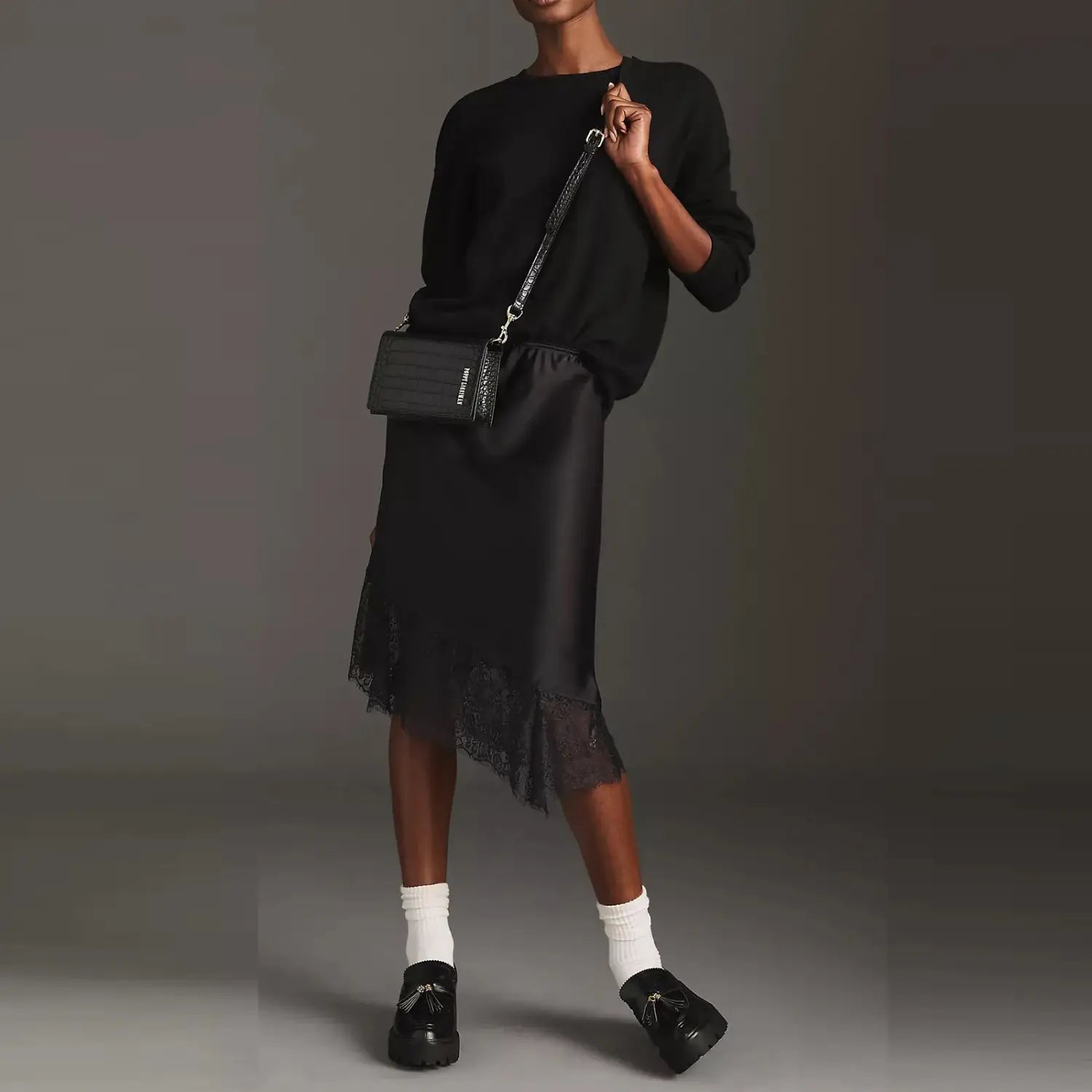 Eine schwarze gesteppte Leder-Crossbody-Tasche mit silbernem Kettenriemen, neben einem Spitzen-Satin-Midirock gezeigt.