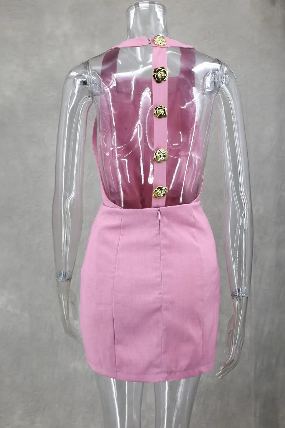 Robe mini rose en satin, sans manches, dos nu, avec un panneau arrière transparent et des boutons floraux d’un ton doré disposés au centre.
