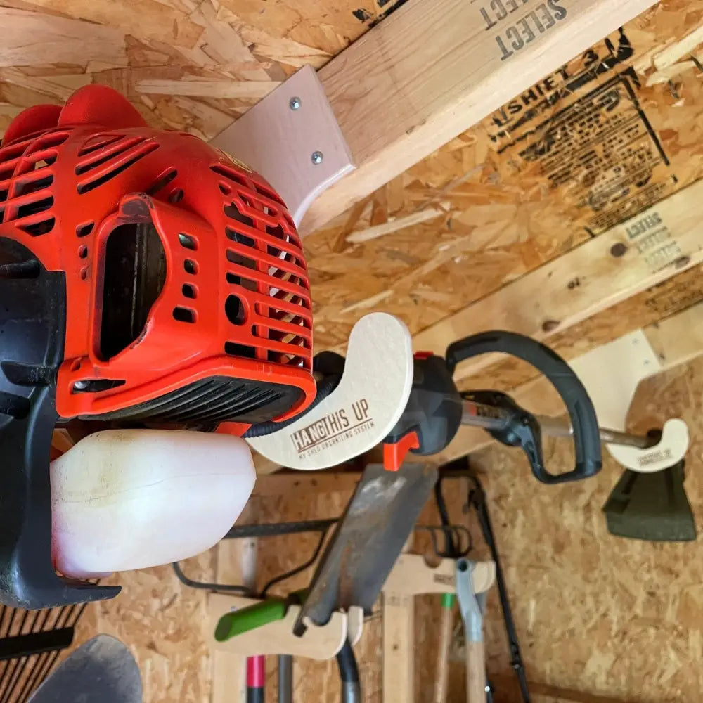 Red and black gas-powered string trimmer with white handle grip mounted on ceiling mounted birch plywood weed wacker rack