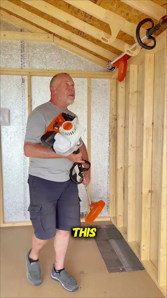 Orange and white handheld string trimmer with black handle and cord stored on ceiling mounted birch plywood weed wacker rack