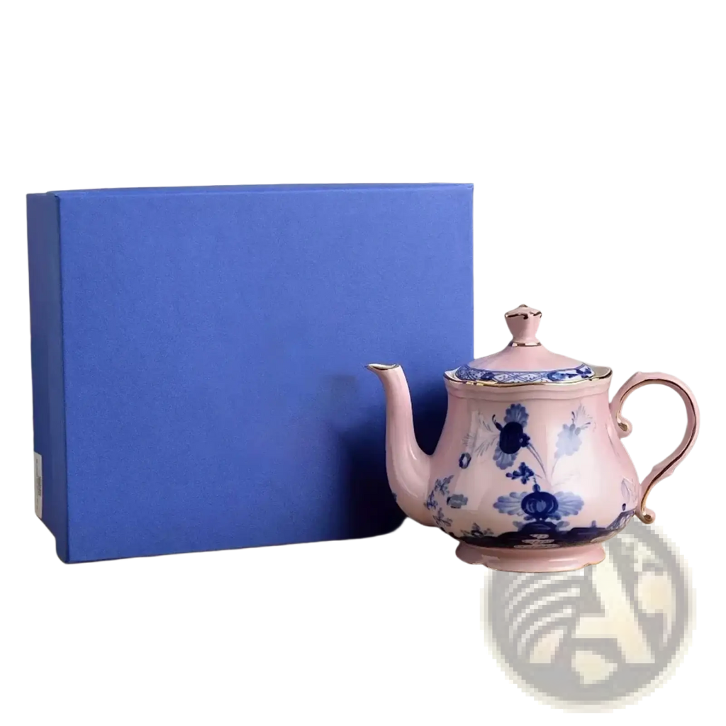A delicate pink porcelain teapot with blue floral patterns and gold trim beside a royal blue gift box for ceramic tableware