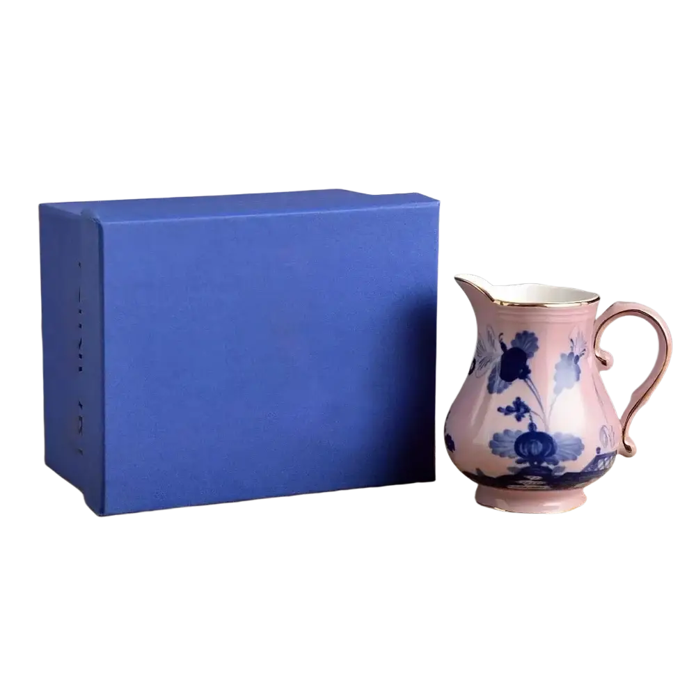 A delicate pink ceramic pitcher with blue floral patterns and gold rim beside a royal blue gift box in a ceramic tableware set