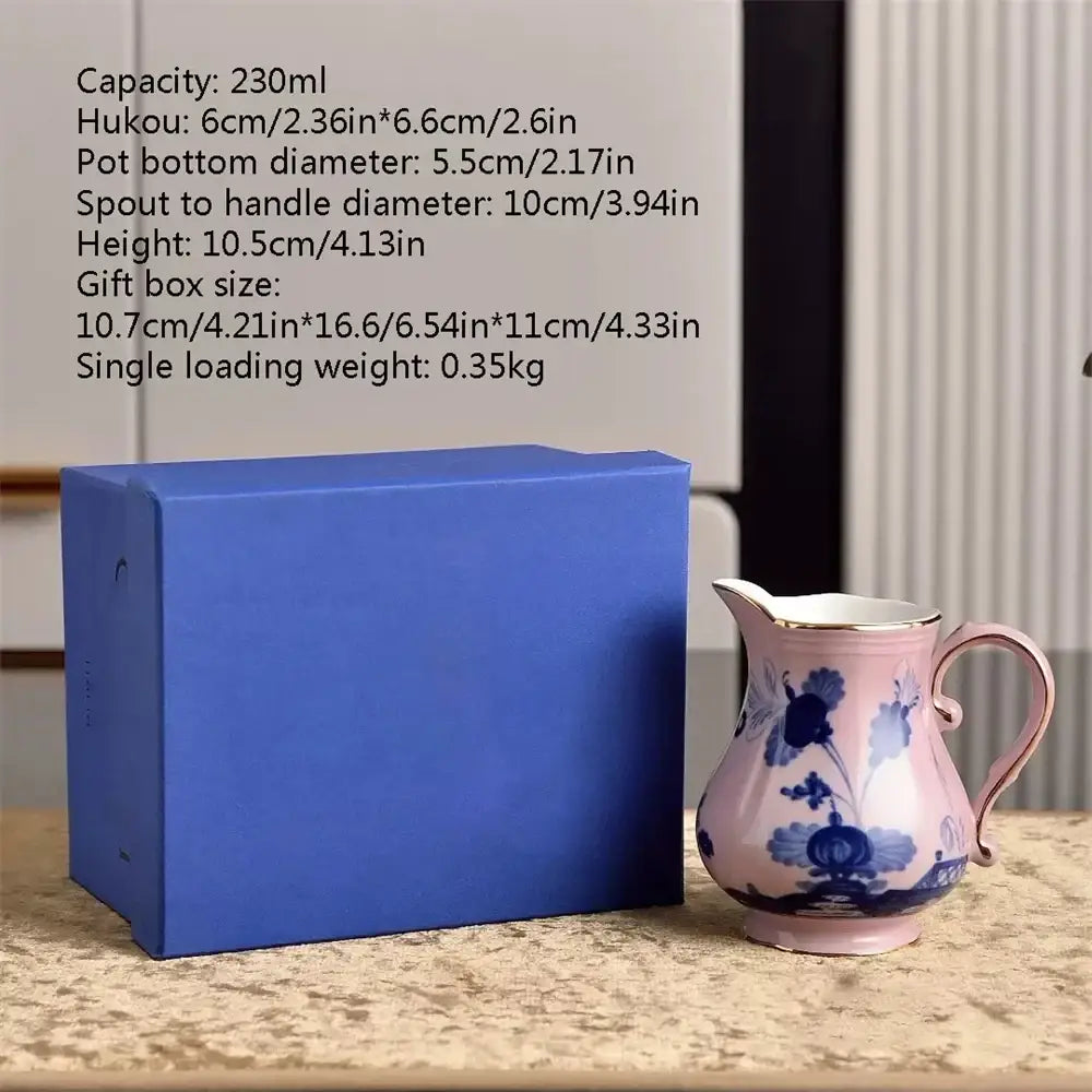 A delicate pink ceramic pitcher with blue floral patterns and gold trim beside its royal blue gift box from the ceramic floral tableware set