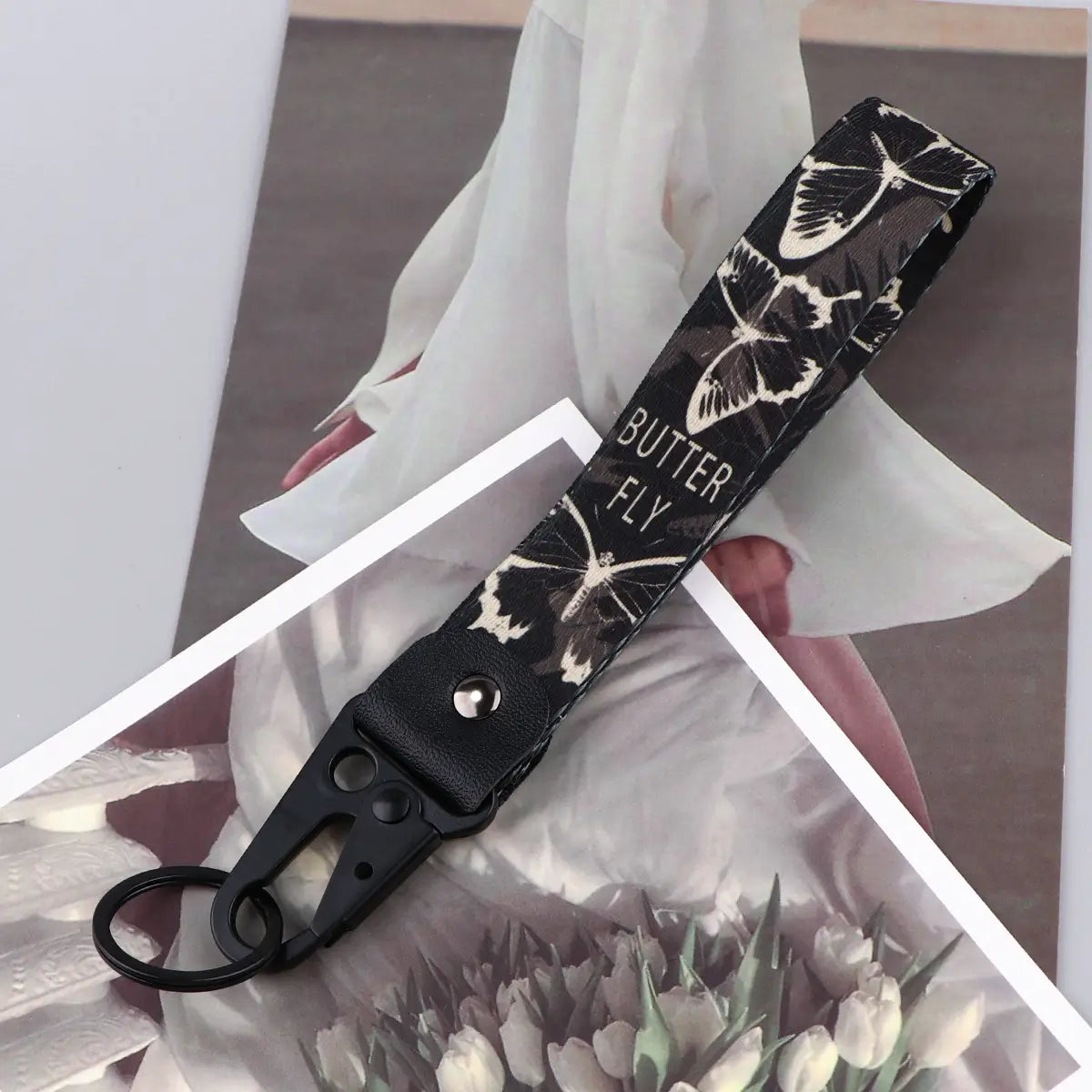 Black wrist strap keychain with white butterfly print and butter fly text featuring a black leather tab and metal carabiner for secure attachment.