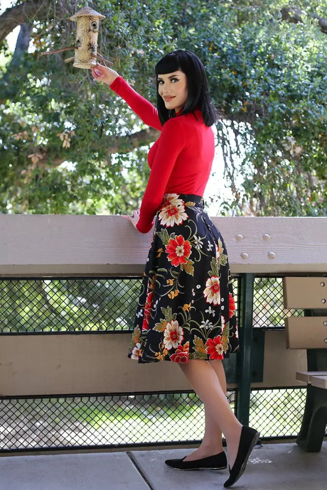 A woman in a red top holds a wooden bird feeder wearing a Charlotte black floral 1950s vintage style circle skirt