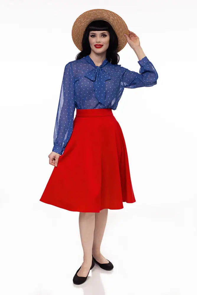 A woman wears a vintage-inspired blue sheer polka dot blouse with a bow and a red Charlotte high waisted circle skirt with pockets