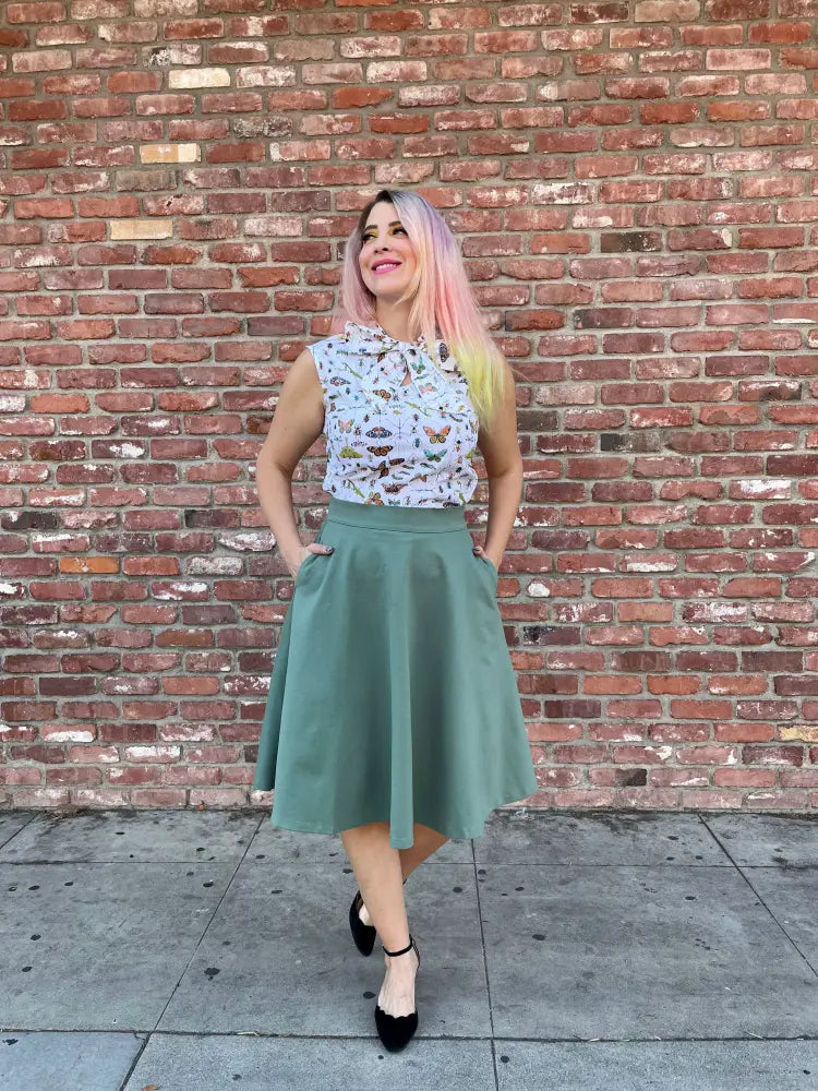 A woman wears a sleeveless butterfly print blouse with a muted green charlotte sage plus size vintage a line circle skirt