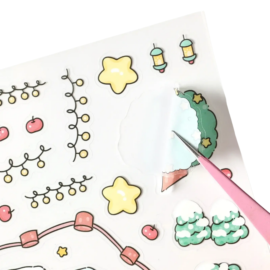 Pink tweezers lifting a green and white snow-covered wreath sticker from a christmas cartoon sticker set sheet