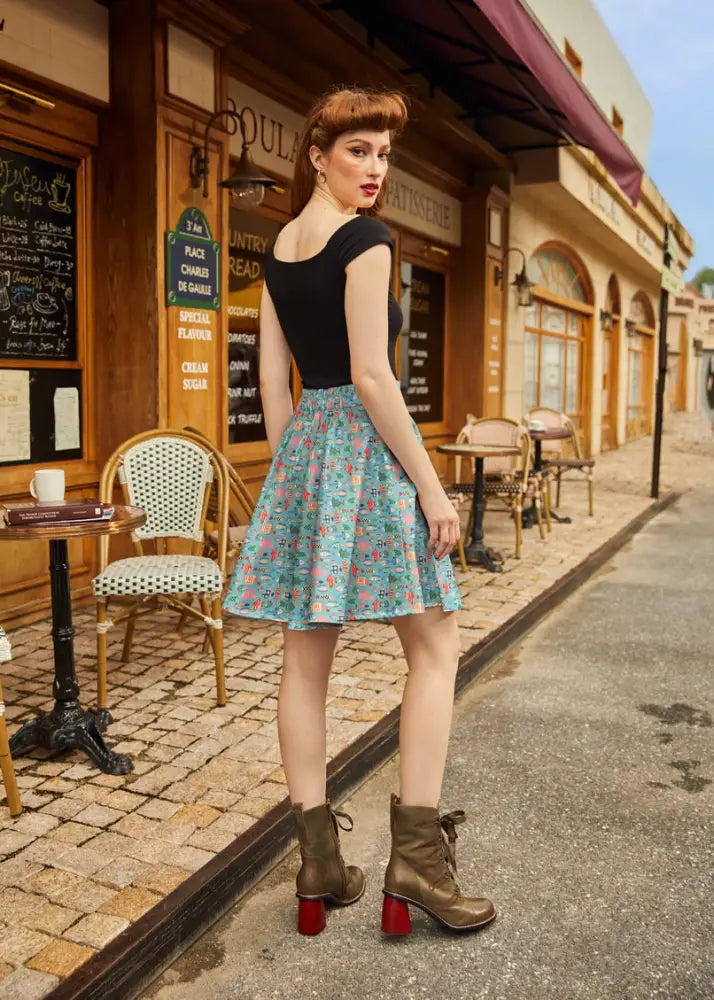 A woman wears a teal colorful fish print vintage a line skater skirt with a black top and brown heeled boots