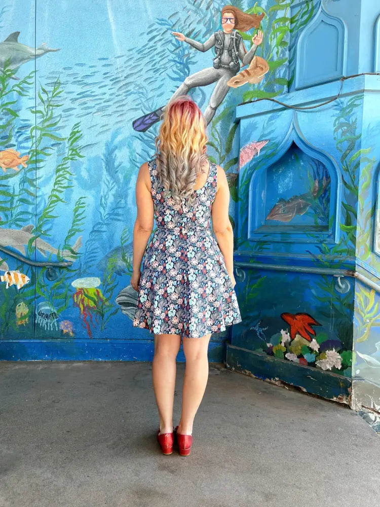 A woman wears a coral floral sleeveless skater dress with pockets and red shoes before an underwater mural