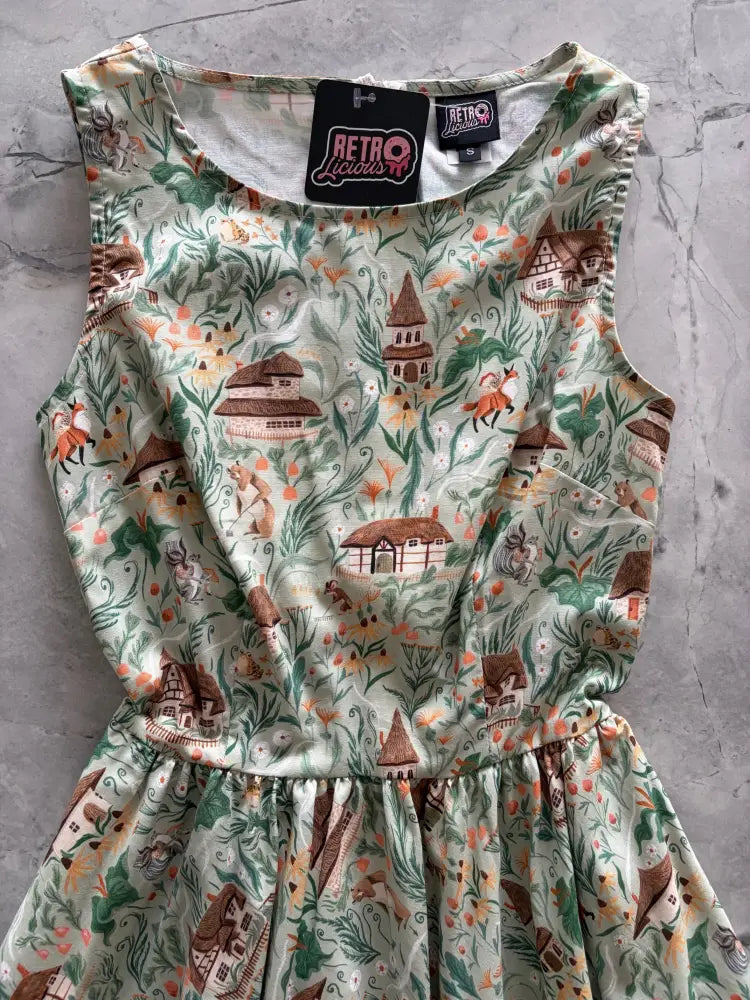 A sleeveless light green cotton midi dress with whimsical fox and house print in earthy tones called Cottage Tales Green Fox Print