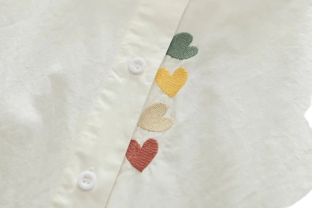 A white cotton button-up blouse with colorful green, yellow, beige, and red embroidered hearts along the placket