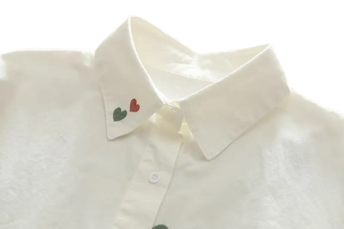 White cotton button up blouse with a crisp collar and delicate red and green heart embroidery near the placket