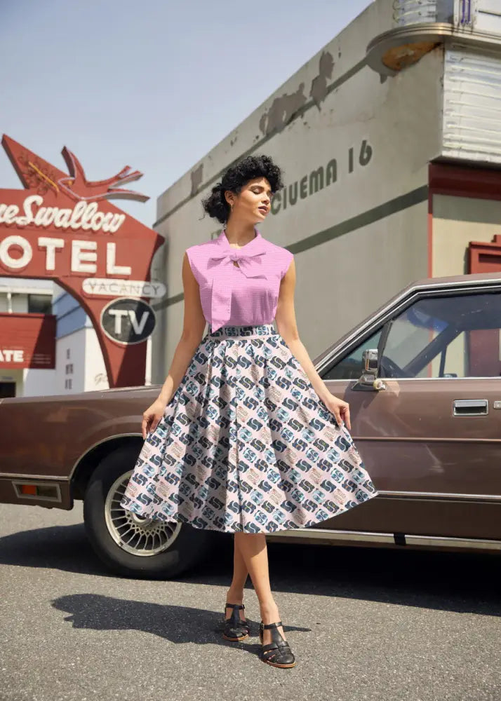 A woman wears a pink sleeveless blouse with a bow and a gray cotton cowgirl boots print Doris retro pleated skirt with pockets