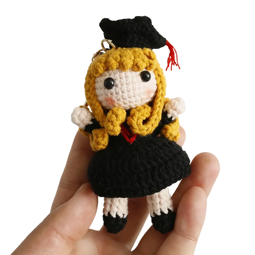 A crocheted graduation doll keychain featuring a blonde-haired figure in black gown and cap with red tassel and white accents